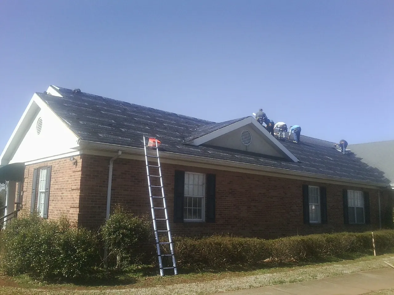 Crew tearing shingles off a brick ranch roof with a ladder set — Emergency Roof Repair work by our crew in Wolflake