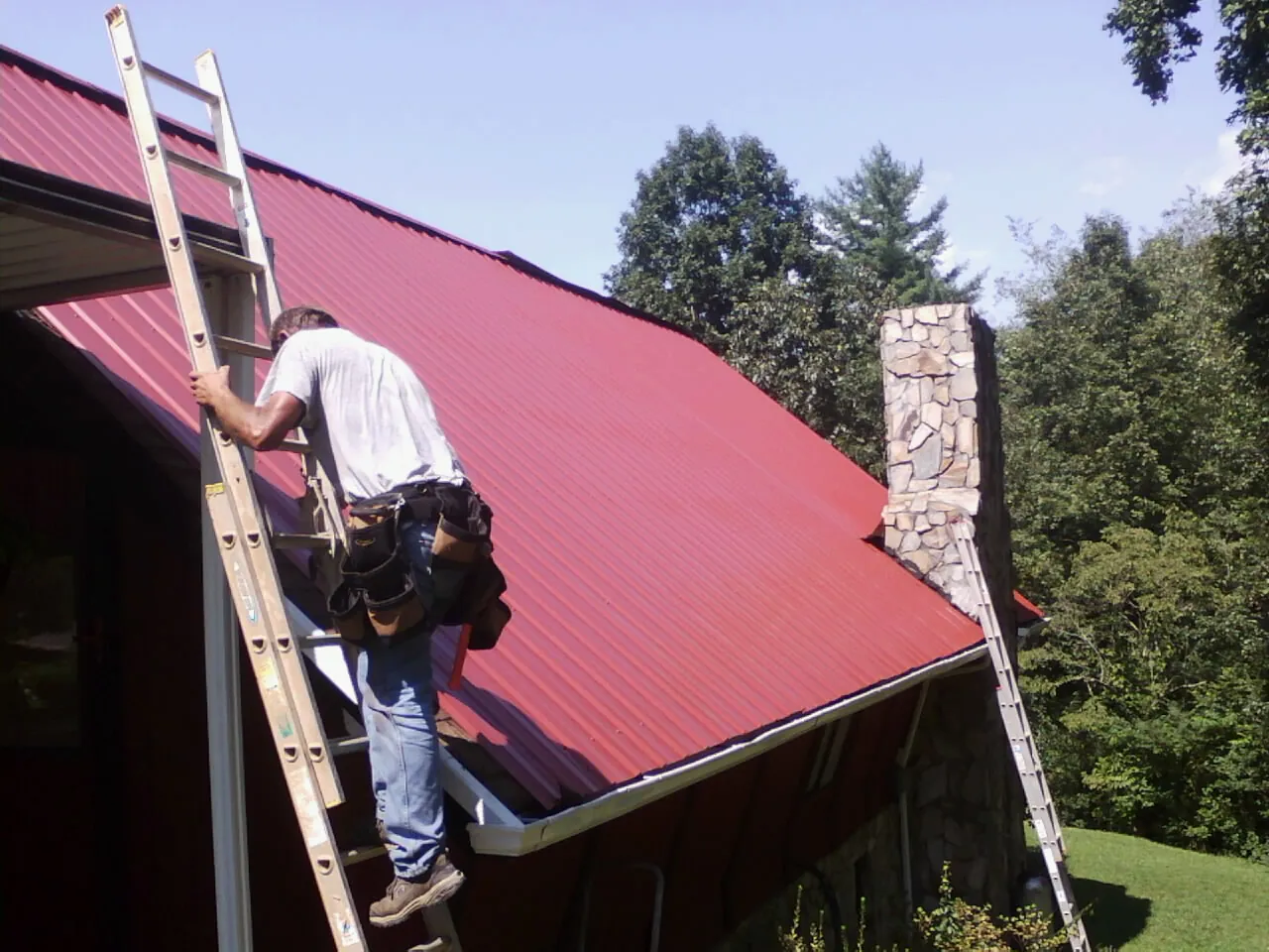 Roofer on a ladder beside a red metal roof and stone chimney — Soffit & Fascia Repair by Roofing Brothers in Wolflake